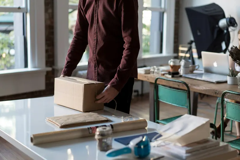 Someone packing an order into a cardboard box at a desk, with a laptop behind.